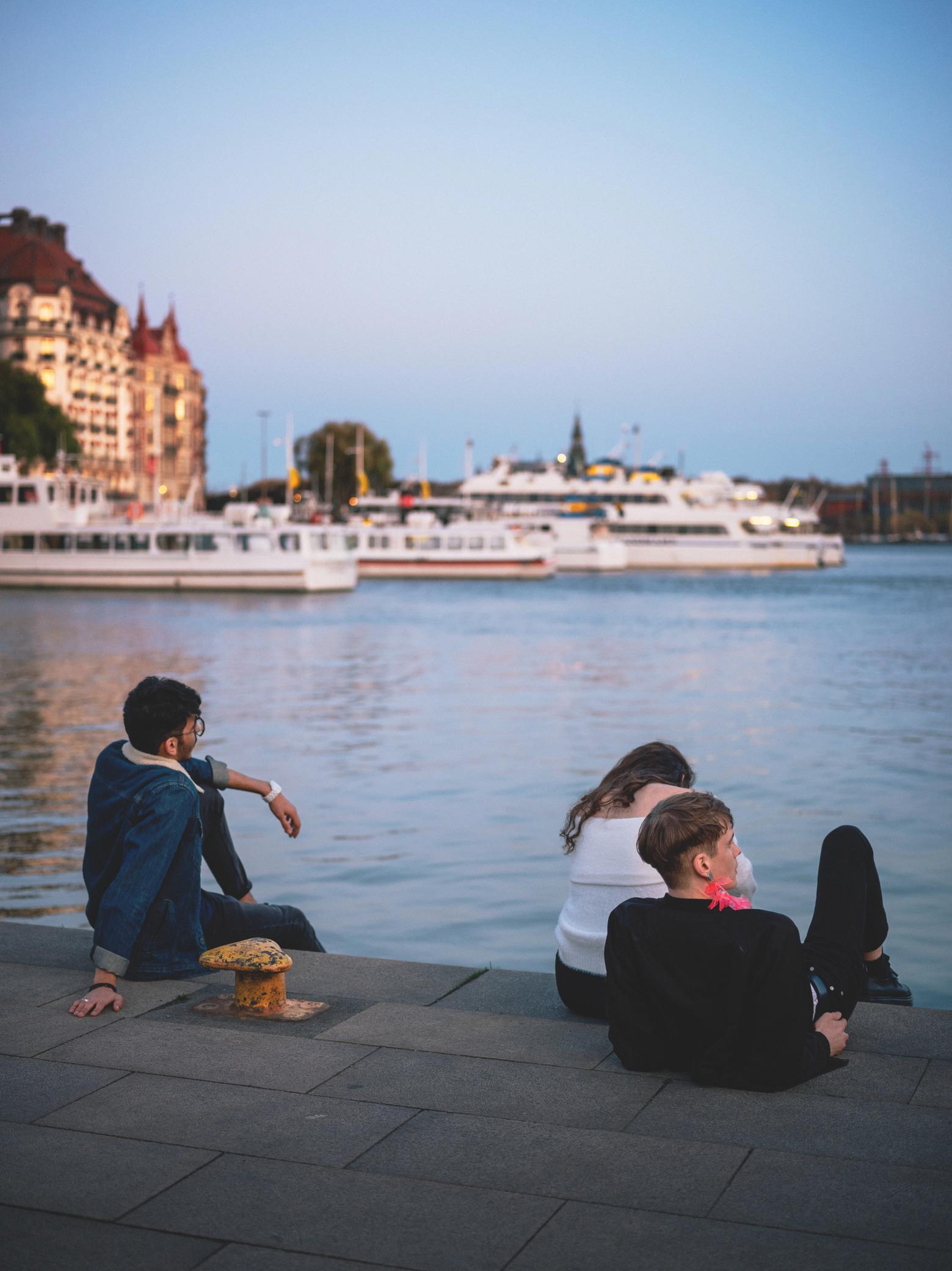 Young professionals relaxing by the Stockholm waterfront at sunset, overlooking ferries and historical architecture, highlighting Sweden’s vibrant urban life