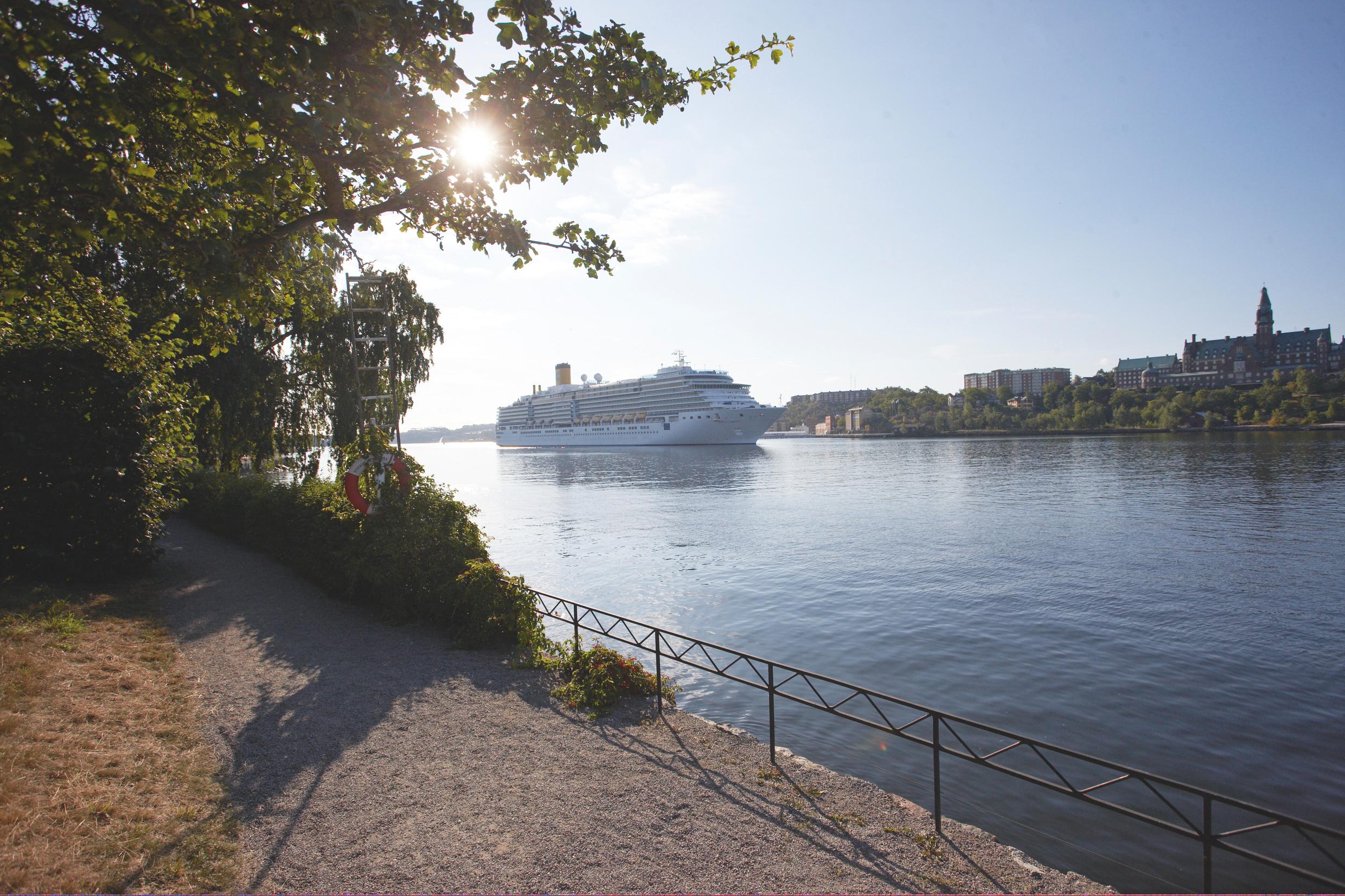 Large cruise ship sailing through Stockholm’s tranquil waterways, surrounded by greenery and cityscape under a bright summer sky.