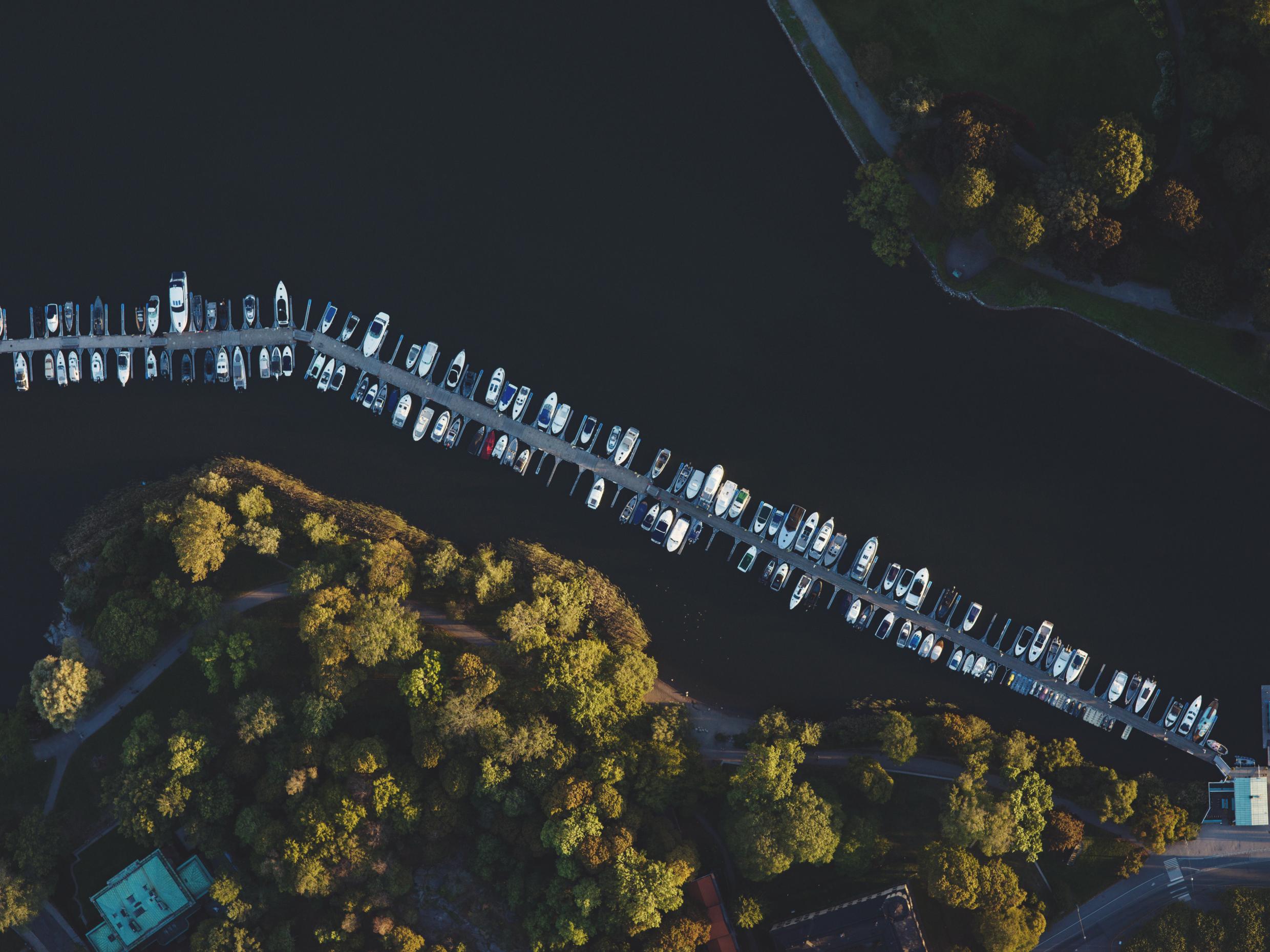 Aerial view of Nobelparken marina in Stockholm, with rows of boats docked along a pier surrounded by lush greenery and calm waters.