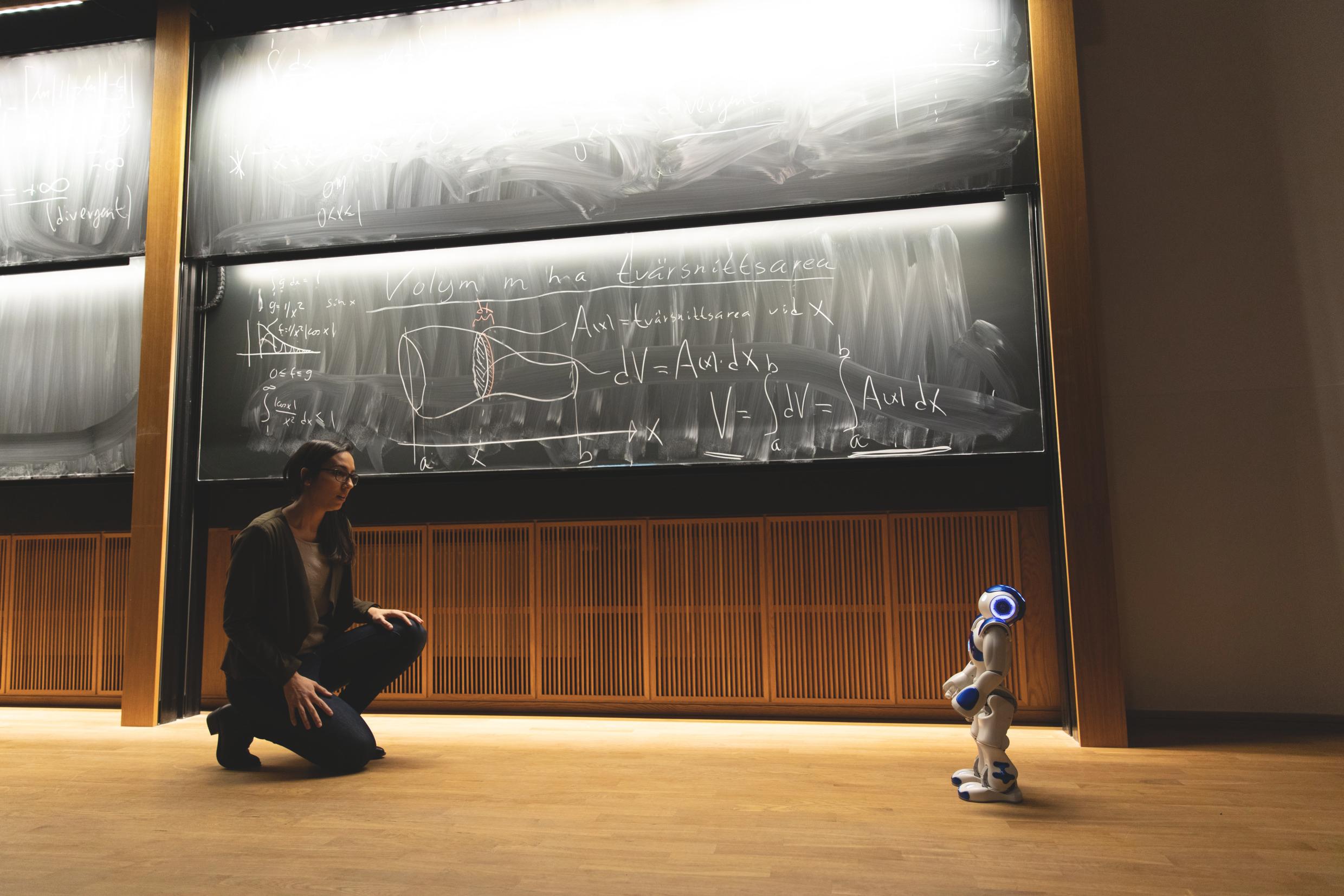 Researcher kneeling in front of a humanoid robot in a lecture hall with chalkboards covered in scientific equations.
