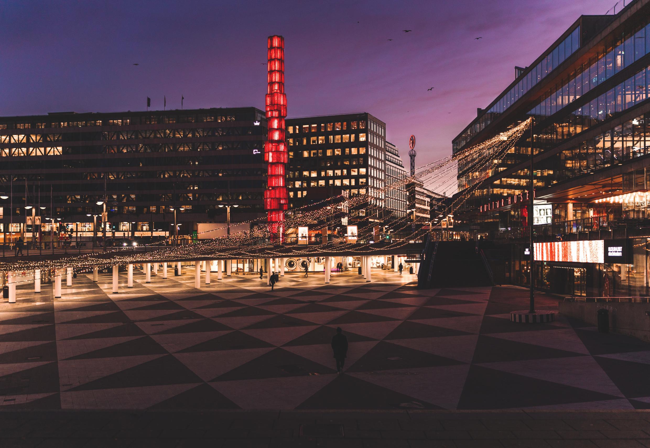 Sergels Torg in Stockholm illuminated at dusk, featuring the iconic red glass column and surrounding modern architecture.